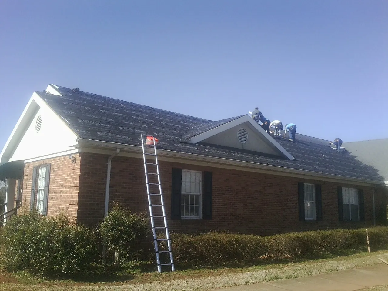 Crew tearing shingles off a brick ranch roof with a ladder set — Metal Roof Repair work by our crew in Paxton