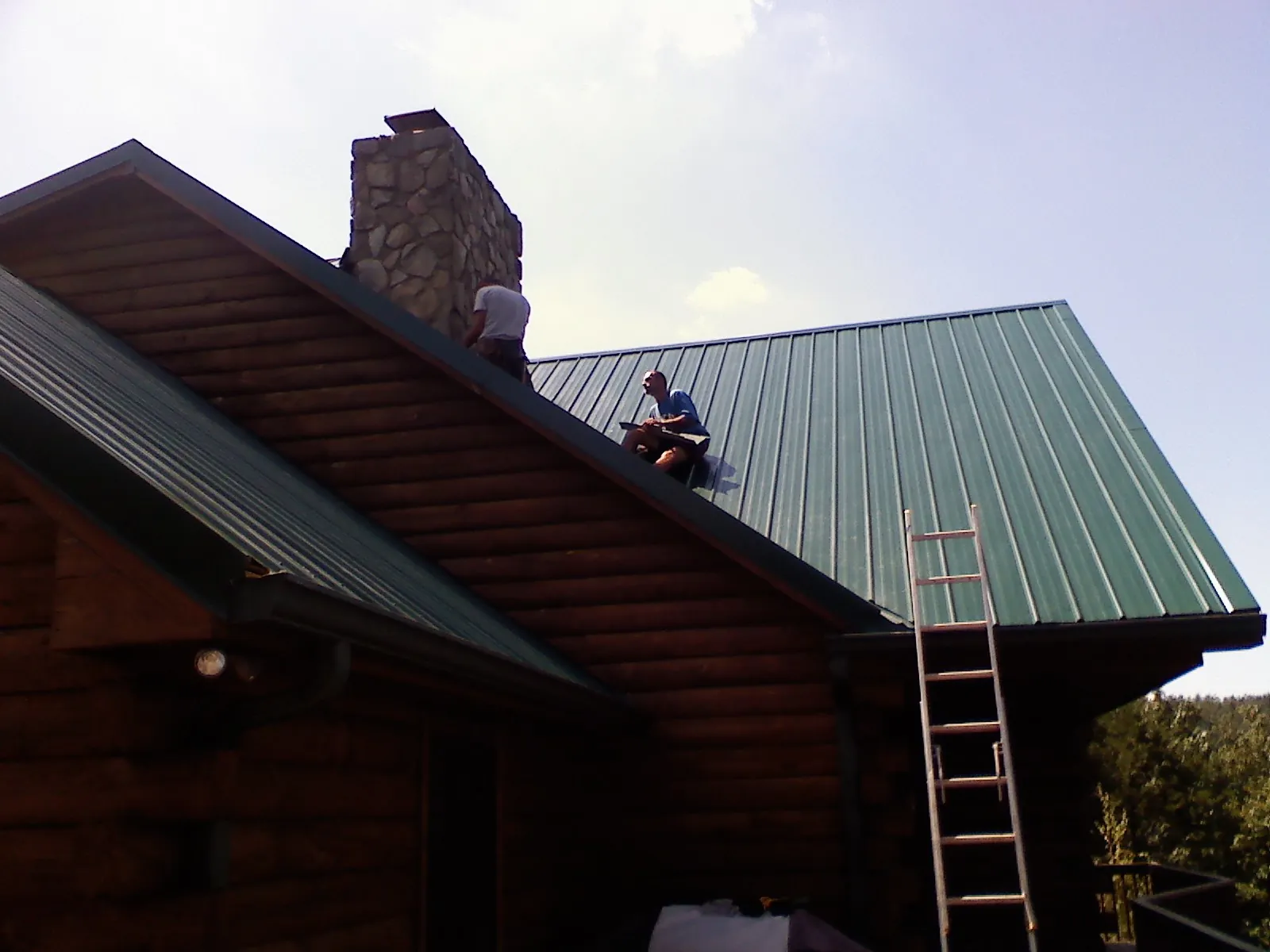 Roofers installing green metal panels beside a stone chimney on a log home in Paxton